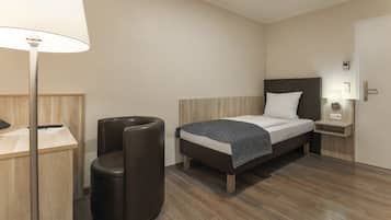 A modern hotel room with a wooden floor, a bed with a white and dark patterned blanket, a desk with a lamp, and a trash bin.