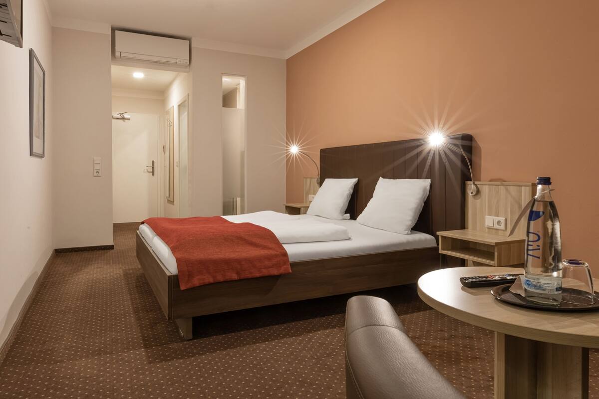 A hotel room with a bed, a bedside table with a bottle and glass, a chair, and a small table with a remote control.