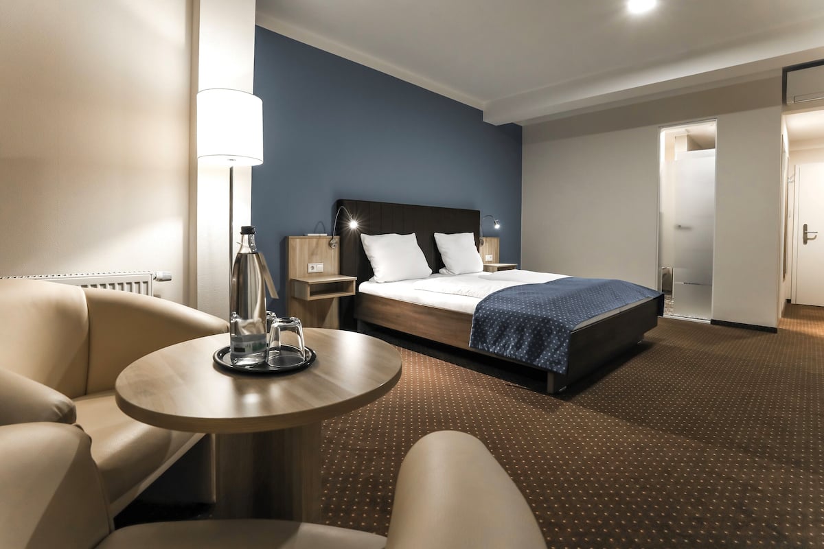 A hotel room with a bed, a round table with a lamp, a chair, and a nightstand.