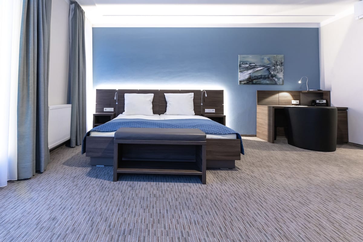 A modern hotel room with a large bed, bedside tables, a desk, and a painting on the wall.