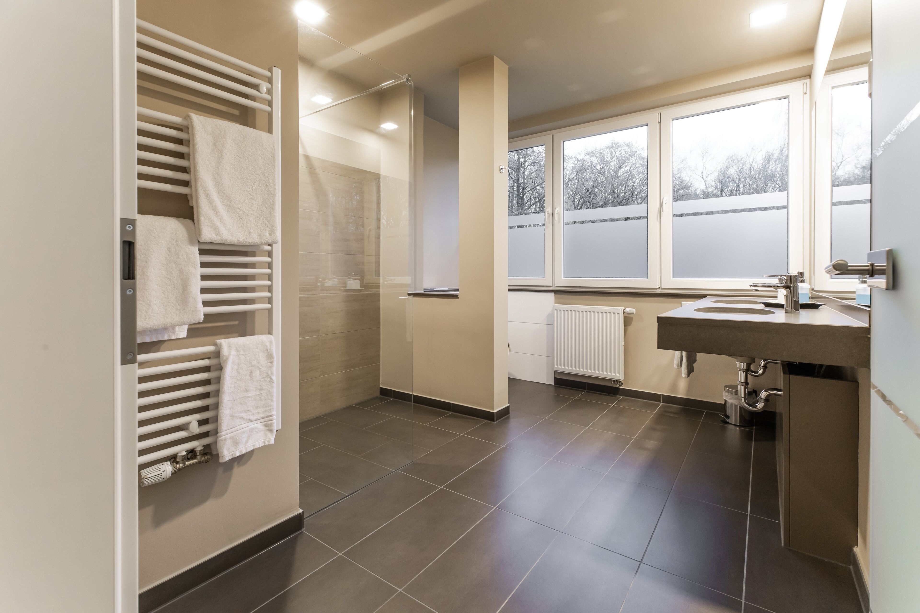 A modern bathroom with a glass shower enclosure, a built-in towel rack, a sink with a mirror, and a window providing a view of the outside.