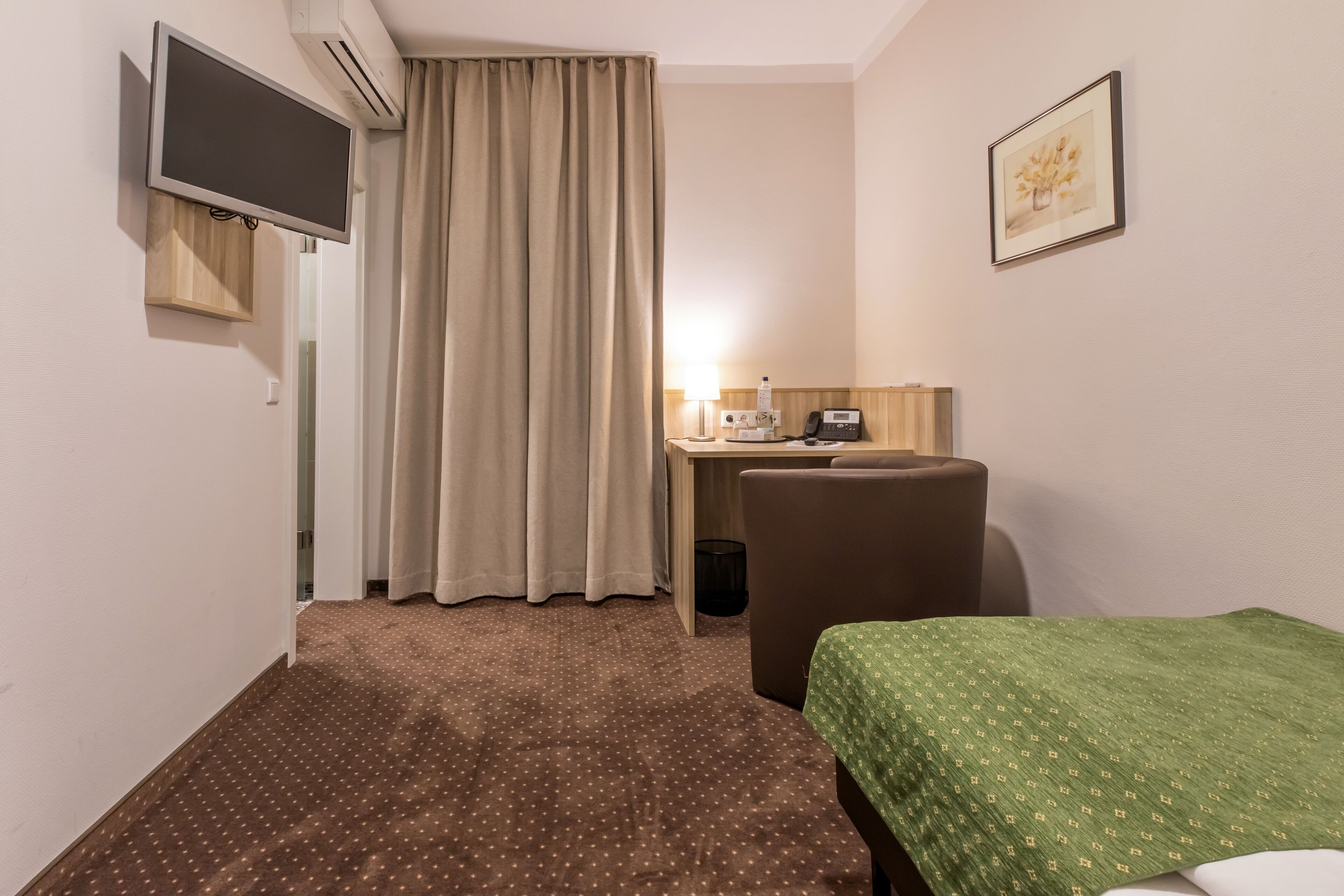 A hotel room with a bed, a desk with a chair, a television mounted on the wall, and a window with curtains.