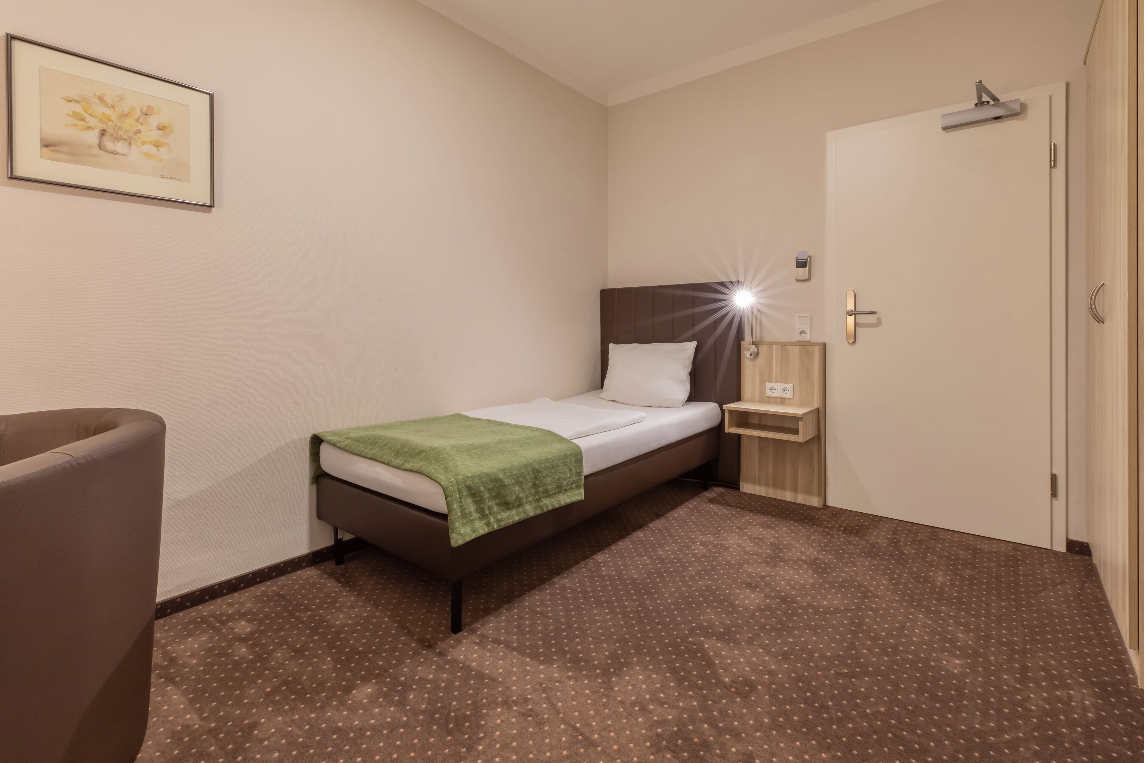 Een kleine, verzorgde slaapkamer met een enkel bed, een nachtkastje, een stoel en een deur.