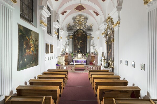 Un interior con hileras de bancas de madera, un altar central y un cuadro grande en la pared izquierda.