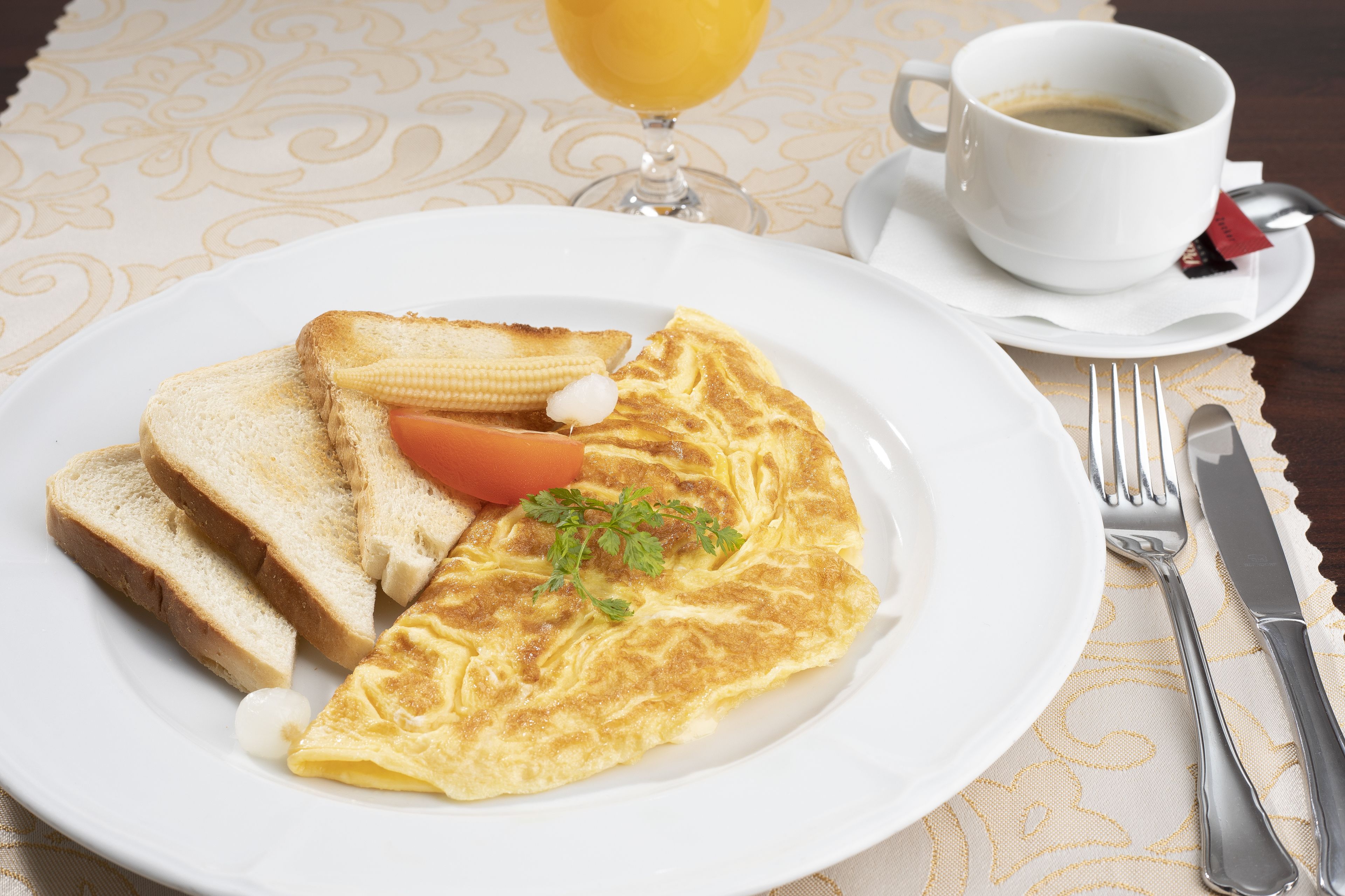 Un plato con tortilla, tostadas y tomate, acompañado de una taza de café y un vaso de zumo de naranja sobre una mesa con posavasos.