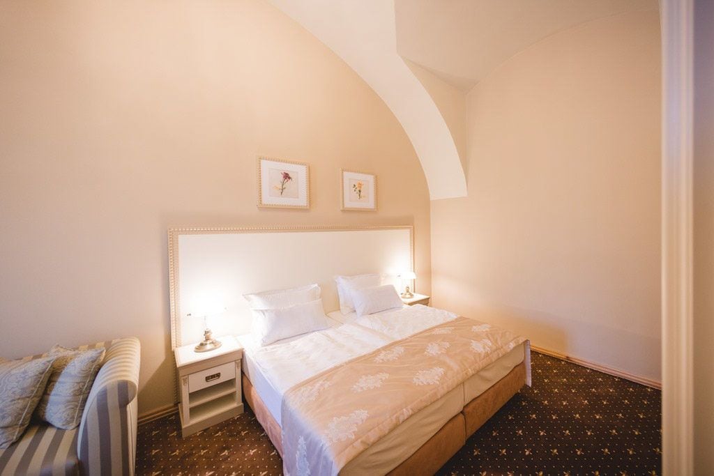 Habitación de hotel con una cama grande, una mesita de noche con lámpara, un pequeño sofá y dos cuadros en la pared.