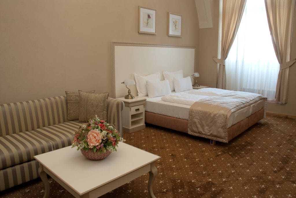 A hotel room with a bed, a sofa, a nightstand, a table with a floral centerpiece, and a window with curtains.