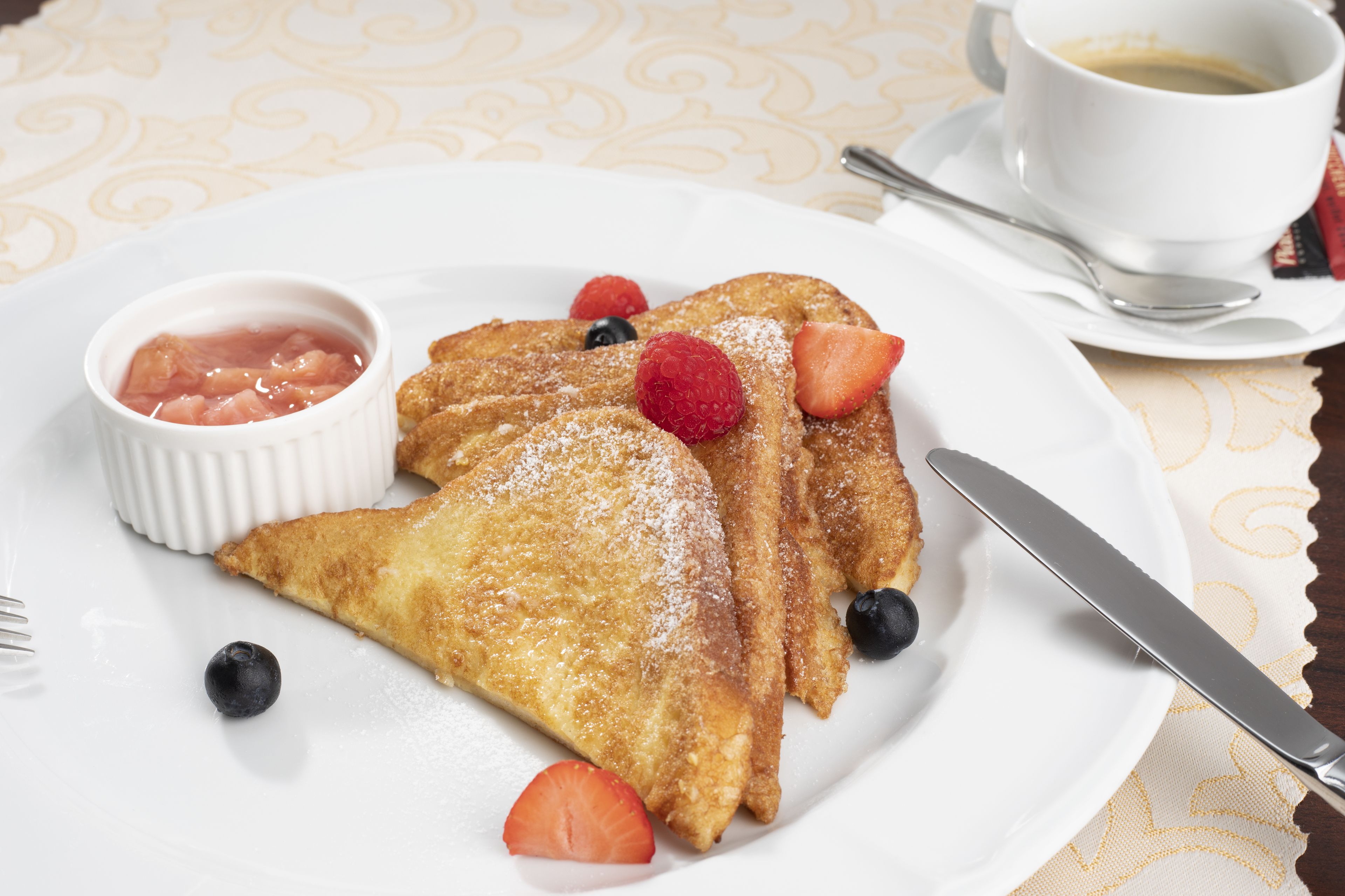 A plate of French toast with strawberries and blueberries, a small bowl of fruit compote, and a cup of coffee on a table with a patterned tablecloth.