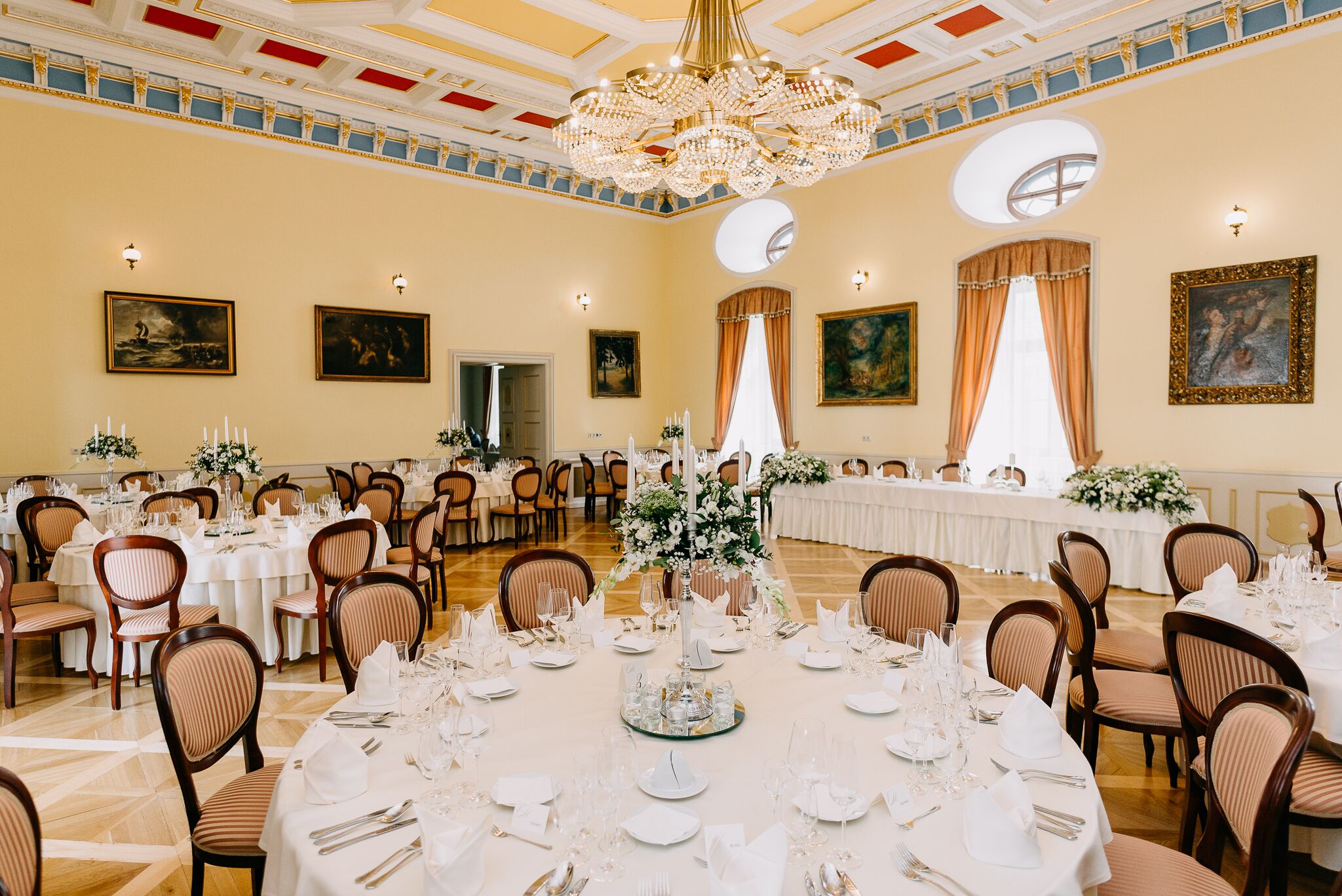 Une salle de banquet avec des tables rondes dressées pour un événement formel, agrémentée de lustres et de tableaux encadrés aux murs.