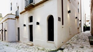 Una stretta via acciottolata, affiancata da edifici con facciate bianche, portali ad arco e piccoli balconi.
