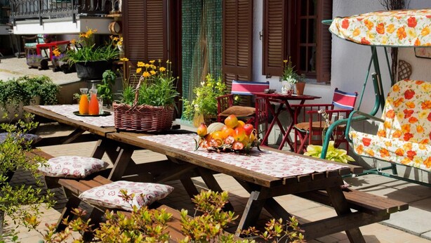 Eine Terrasse im Freien mit einem Holztisch, gedeckt für ein Essen, umgeben von Topfpflanzen und einem Blumenschirm.