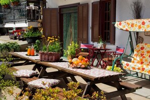 Une terrasse extérieure avec une table en bois dressée pour le repas, entourée de plantes en pot et d’un parasol orné de motifs floraux.