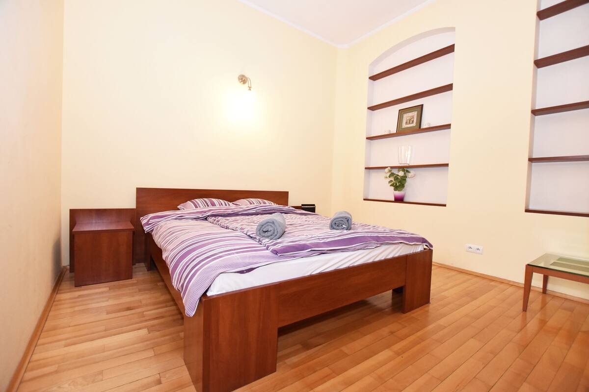 A bedroom with a wooden bed, a nightstand, a bedside table, a wall-mounted shelf with a plant, and a window with blinds.