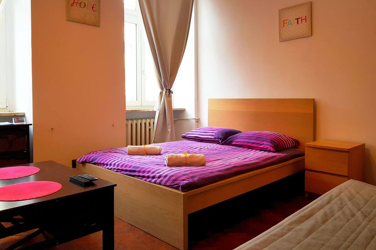 A bedroom with a bed, a nightstand, a desk, and a window with curtains.