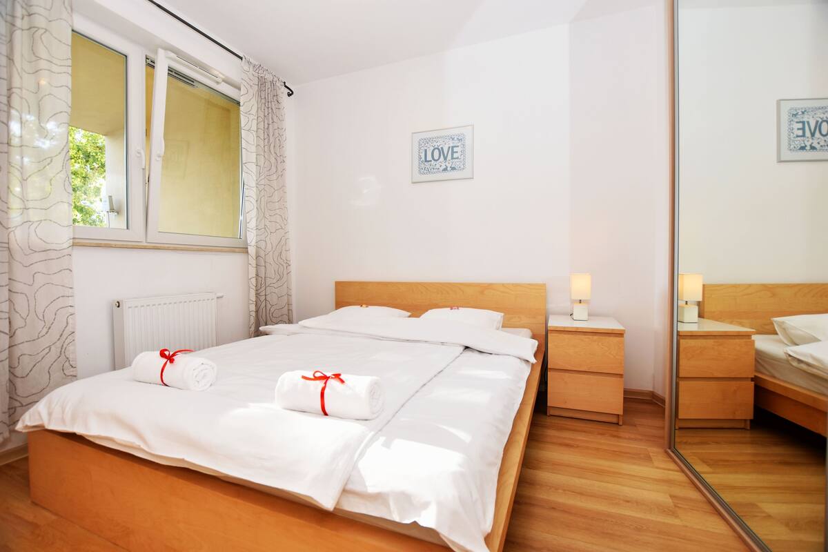 A neatly made bed with white linens and two rolled towels tied with red ribbons, a bedside table with a lamp, and a framed 'LOVE' sign on the wall.