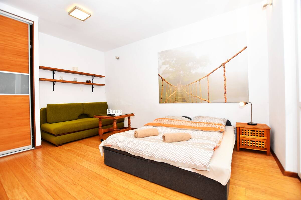 A modern bedroom with a wooden floor, a green sofa, a bed with beige and white bedding, a wooden nightstand, and a hammock bridge painting on the wall.