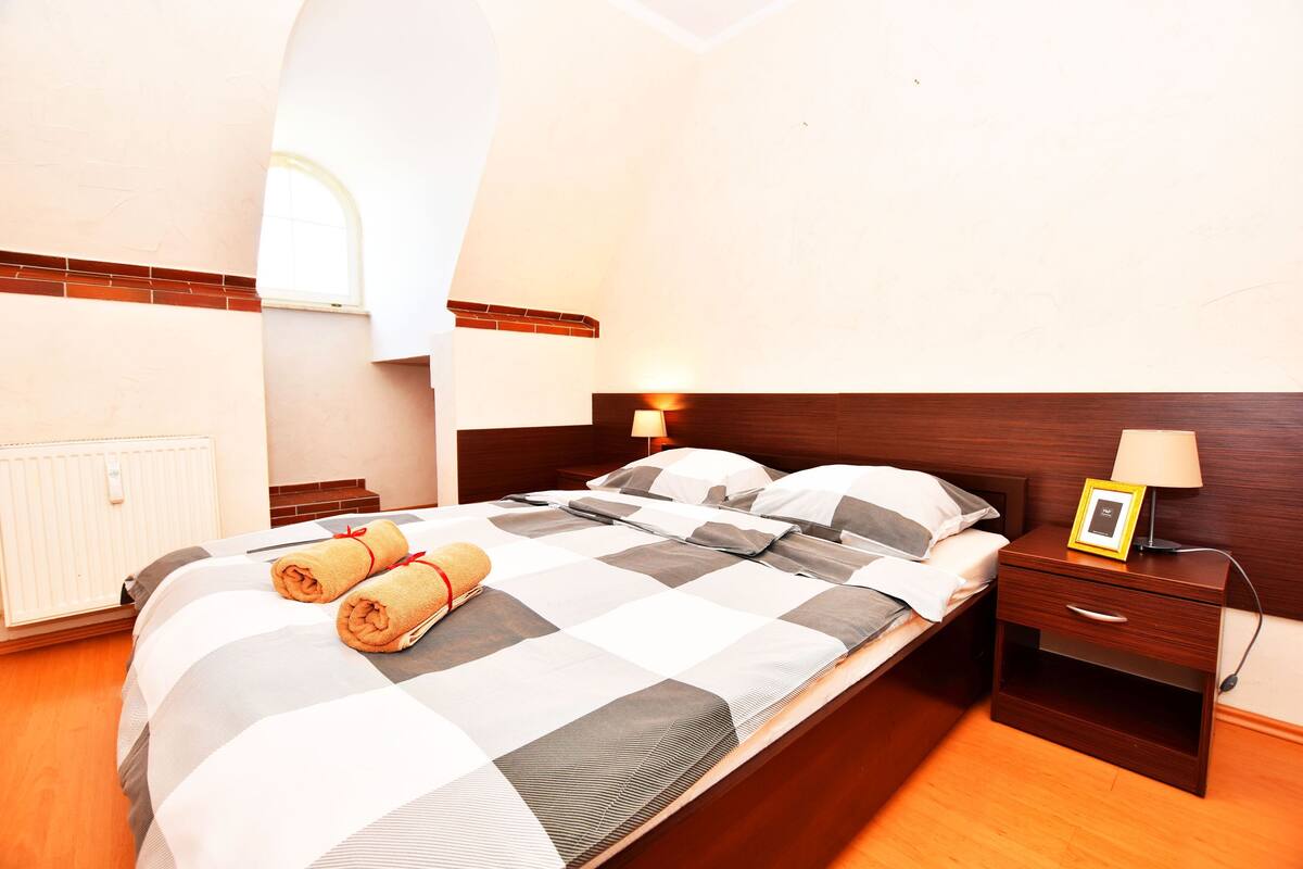 A neatly made bed with a checkered bedspread, two rolled towels, and a bedside table with a lamp and a picture frame.