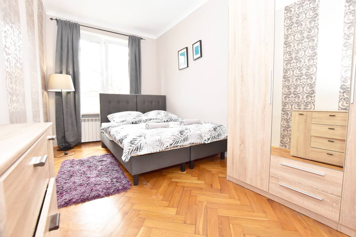 A bedroom with a bed, a dresser, a nightstand, a lamp, and a window with curtains.