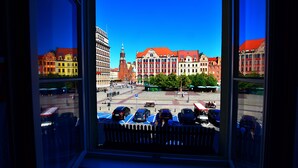 Une vue depuis une fenêtre d’un immeuble, avec un parking en contrebas, où l’on aperçoit plusieurs voitures et un ciel bleu dégagé.