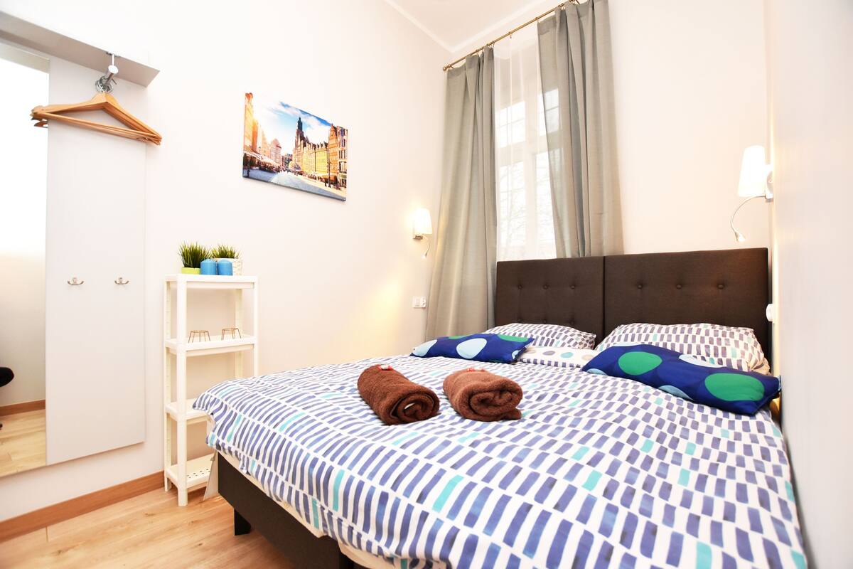 A neatly arranged bedroom with a bed, pillows, a hanging coat rack, a small shelf with a plant, and a framed picture on the wall.