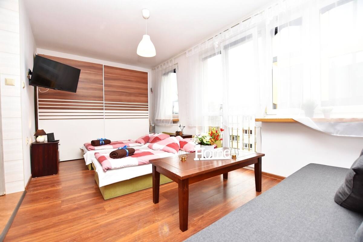 A modern hotel room with a wooden floor, a sofa, a bed with pink and white bedding, a wooden table, and a TV mounted on the wall.