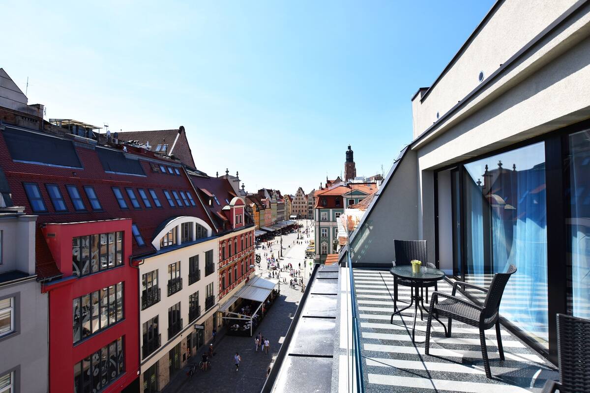A balcony with outdoor seating, overlooking a pedestrian street with historic buildings.