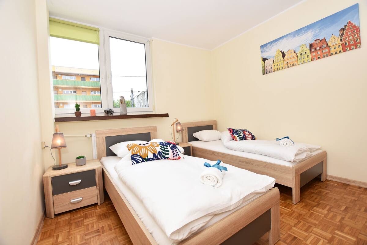 A room with two wooden beds, a nightstand with a lamp, and a window with a view of buildings.
