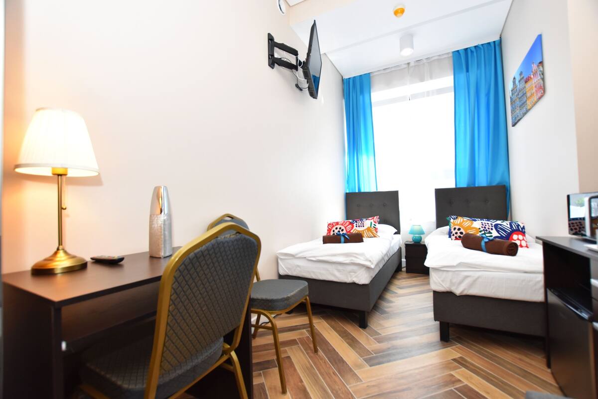 A hotel room with two beds, a desk, a chair, a TV, and a window with blue curtains.