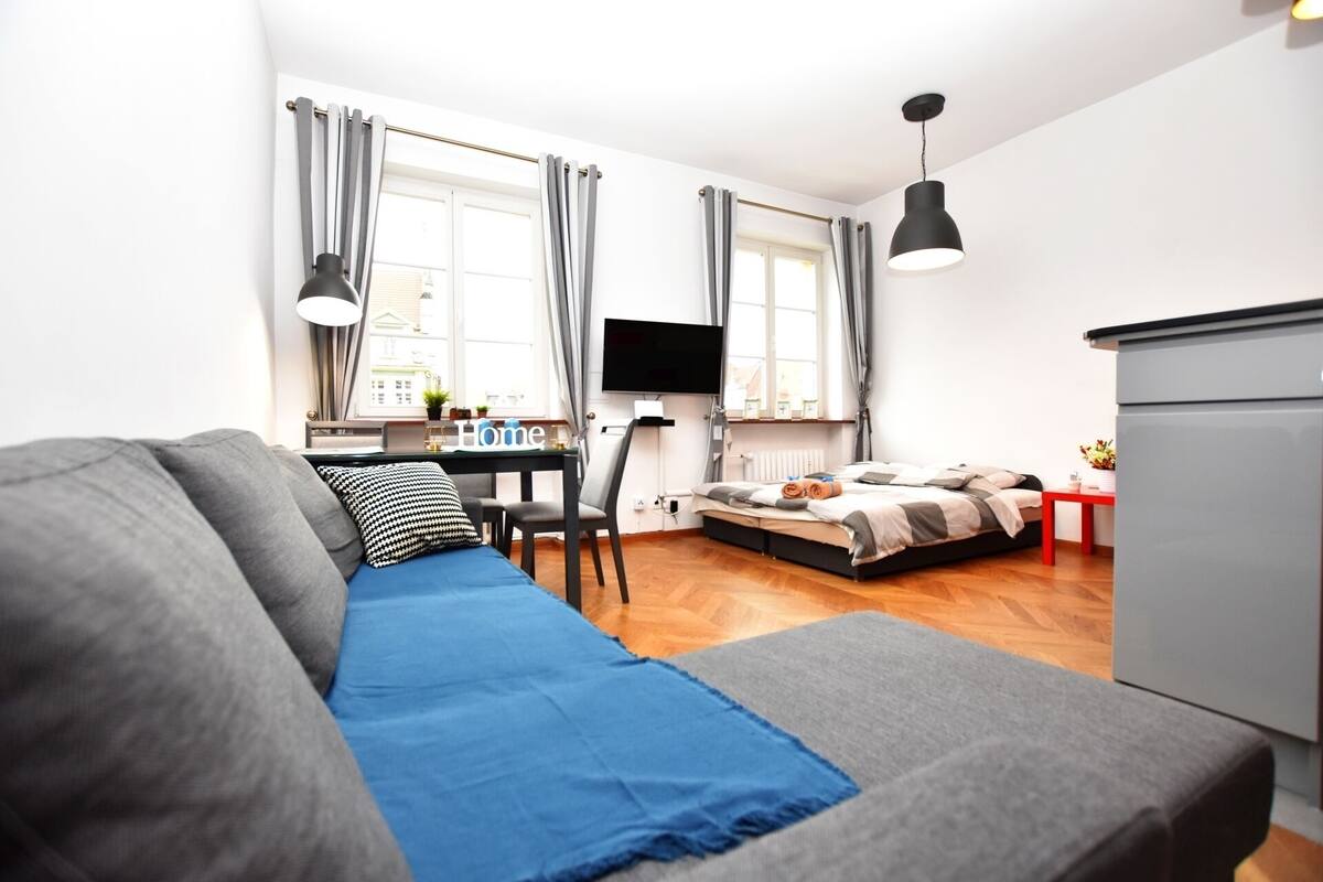 A modern bedroom with a grey sofa, a bed with a grey and white duvet, a small desk with a TV, and a red table with a plant.