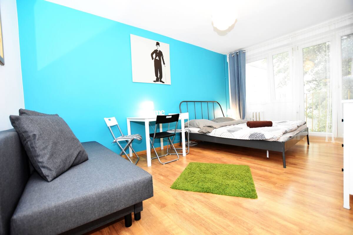 A modern bedroom with a grey sofa, a small desk with a chair, a bed with a green rug, and a large window.