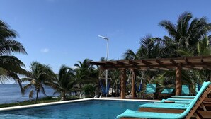 A pool area with lounge chairs and a pergola, surrounded by palm trees and a view of the ocean. - Aquastar By RT Vacation Rentals (Mahahual)