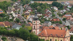 Uma cidade com uma igreja notável, com uma torre do relógio, rodeada por casas residenciais de telhados coloridos e um cemitério ao primeiro plano.