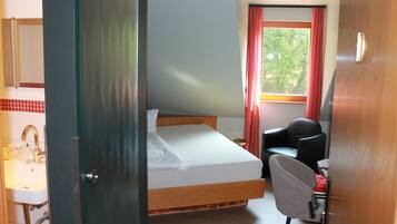 A hotel room with a bed, a desk, a chair, a window with curtains, and a bathroom with a sink and mirror.