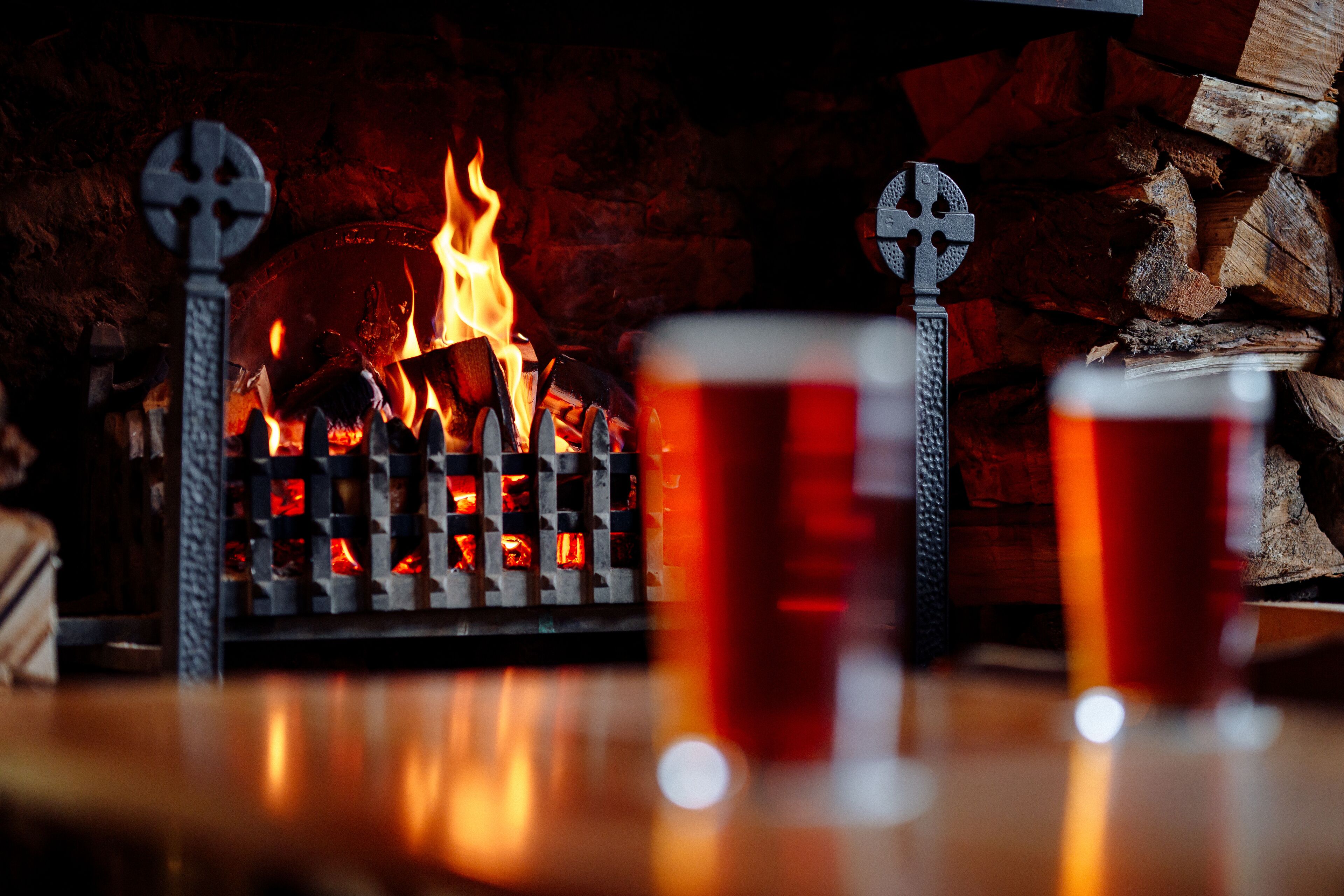A fireplace with logs and a fire, two metal crosses, and two glasses of amber liquid.