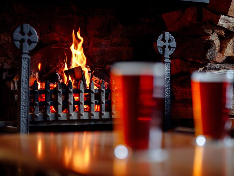 Ein Kamin mit Holz und Feuer, zwei Metallkreuze und zwei Gläser bernsteinfarbener Flüssigkeit.