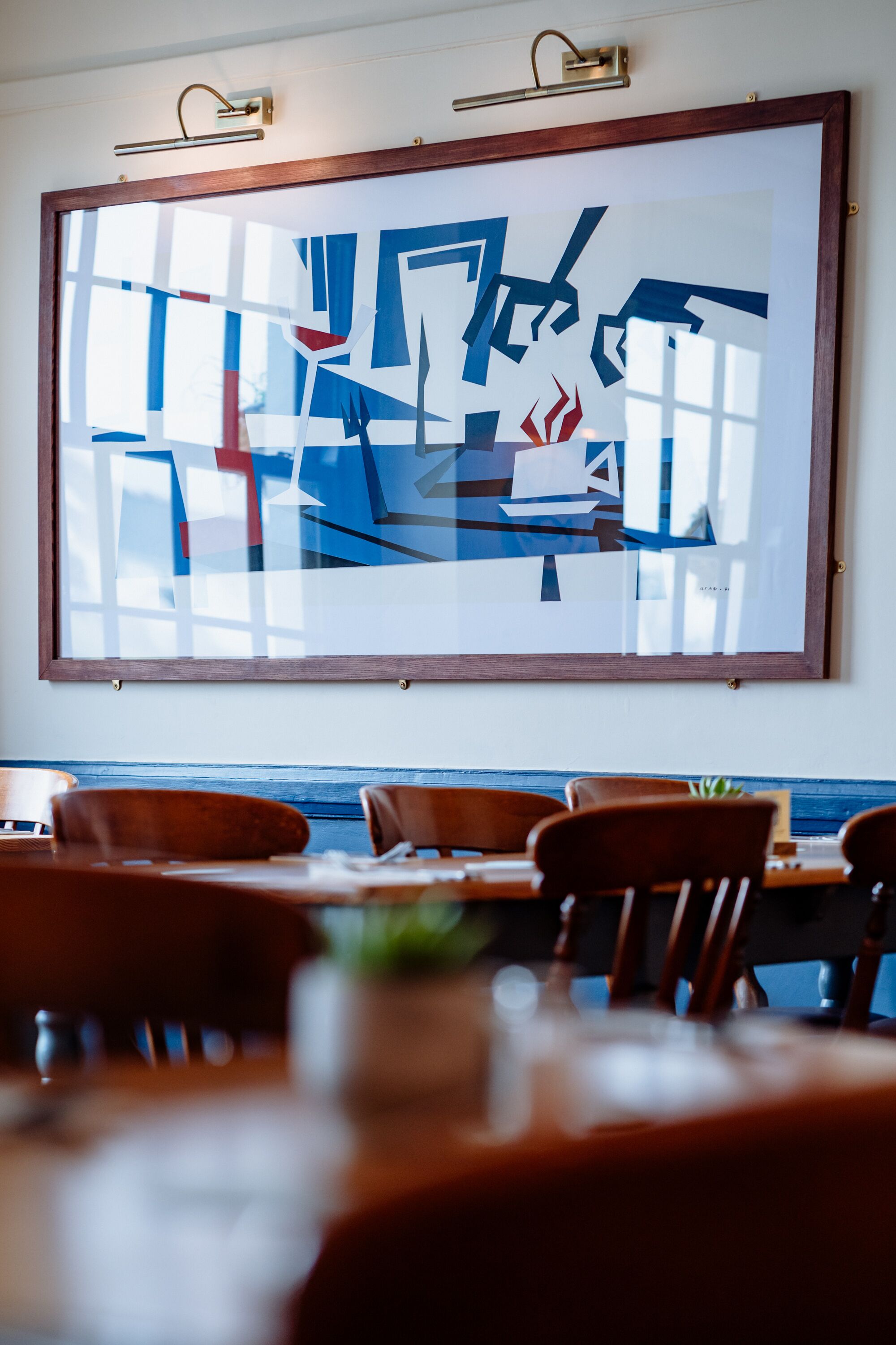 A framed abstract painting on a wall above a dining area with wooden chairs and tables.