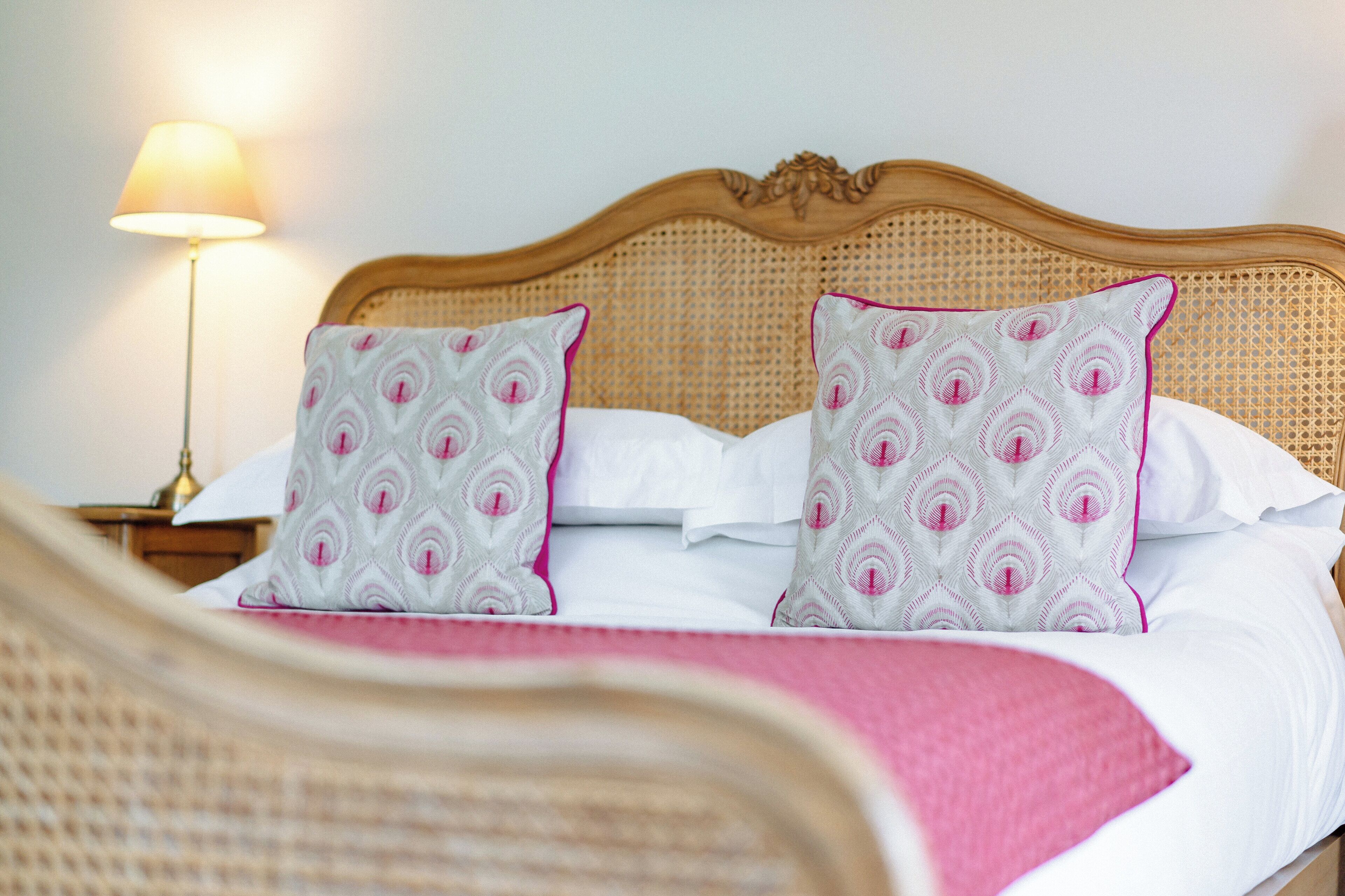 A bed with a woven headboard, two patterned pillows, a white bedspread, and a bedside lamp.