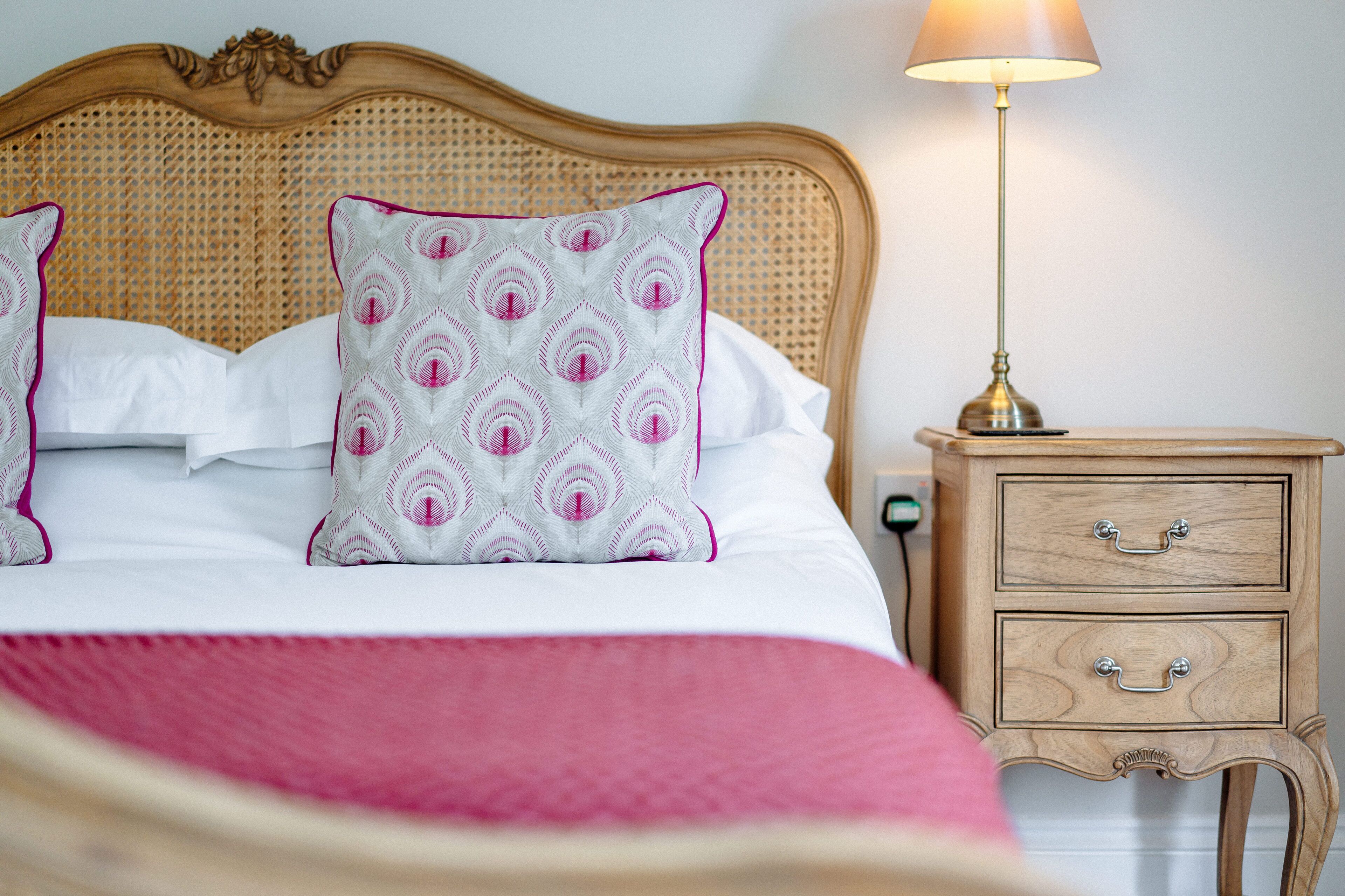 A neatly made bed with a white and pink patterned pillow, a wooden nightstand with drawers, and a bedside lamp.