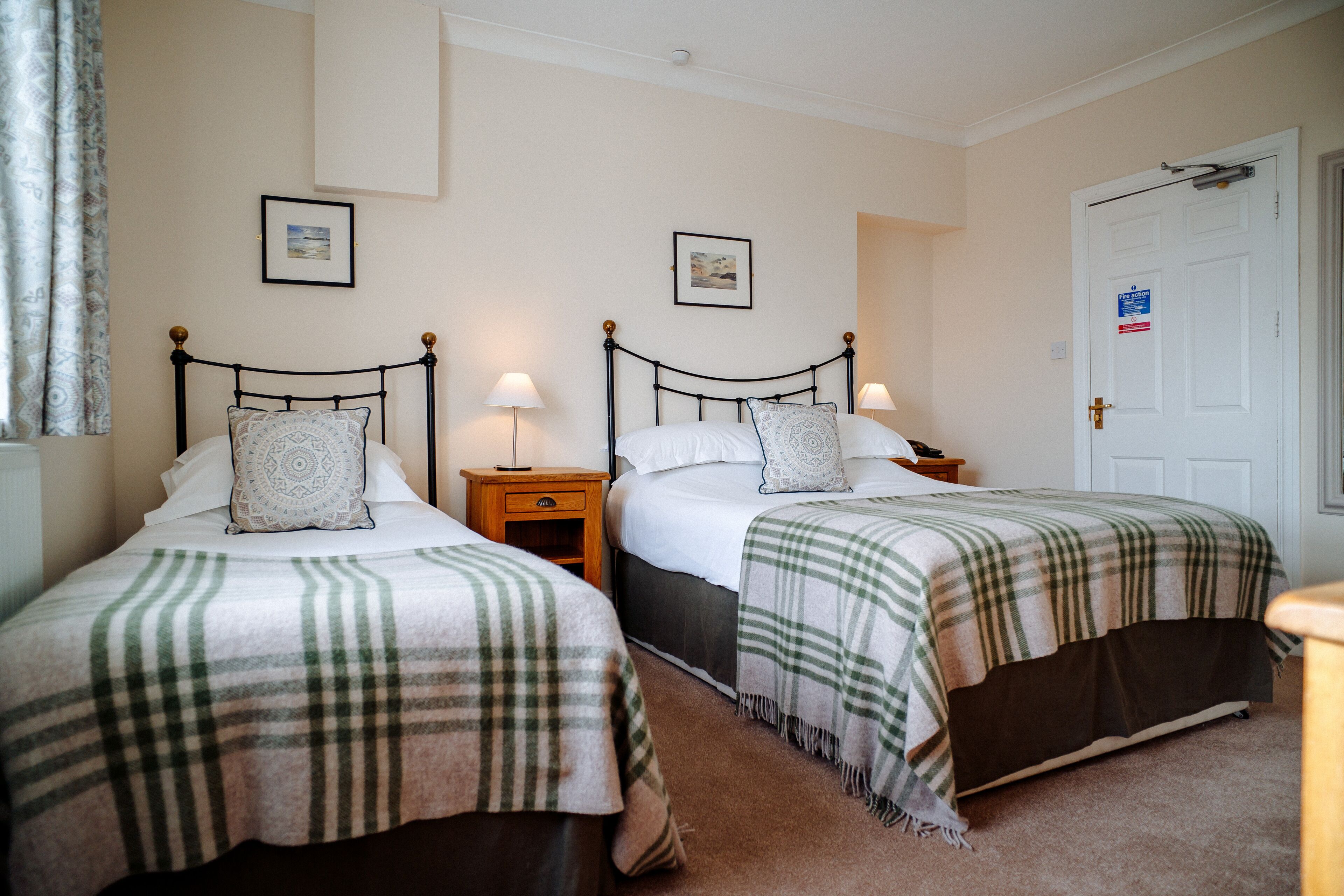 A bedroom with two beds, each with a plaid blanket, bedside tables with lamps, and framed pictures on the wall.