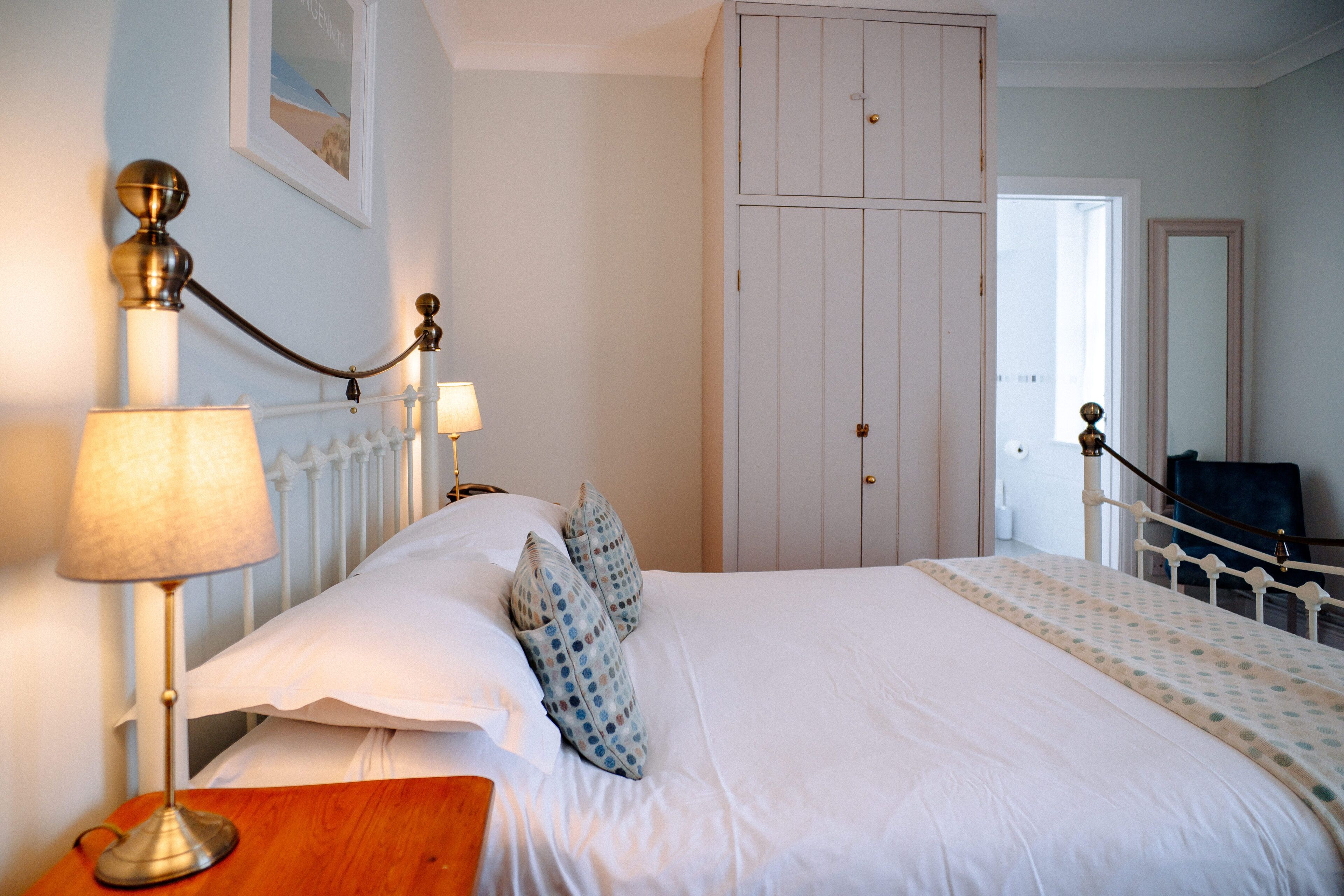 A neatly made bed with white linens and patterned pillows, a bedside table with a lamp, and a white wardrobe in the background.