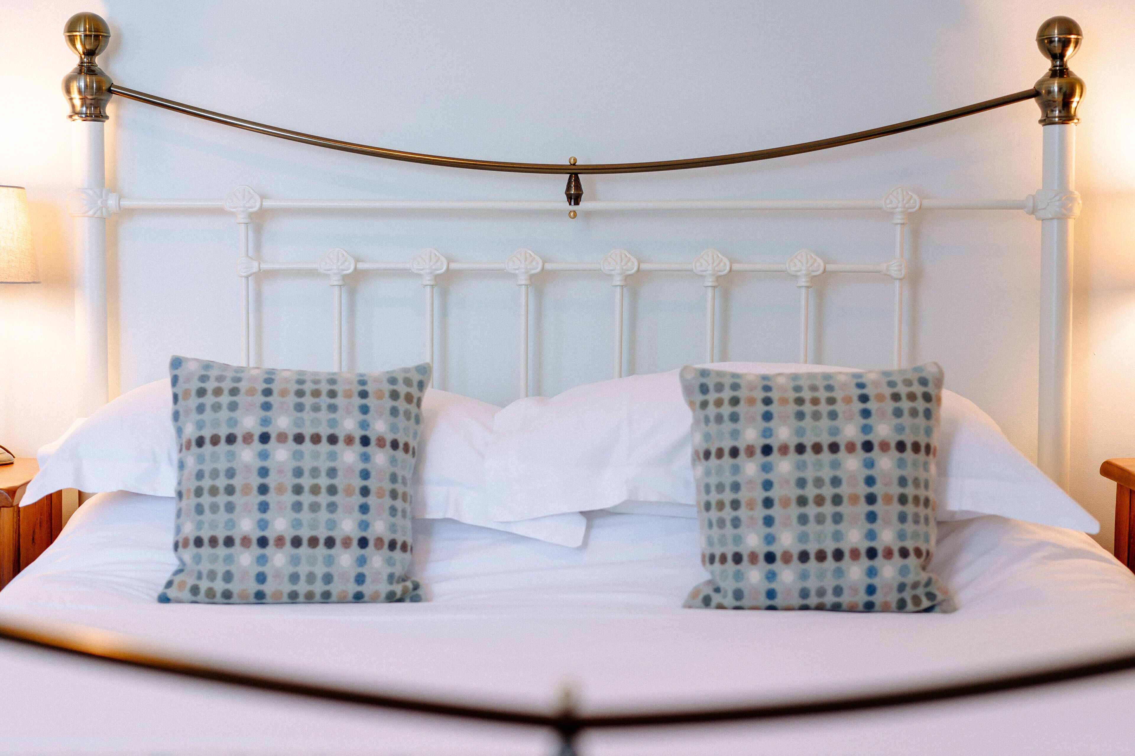 A neatly made bed with white bedding and two patterned pillows, a white headboard, and a brass rod across the top.