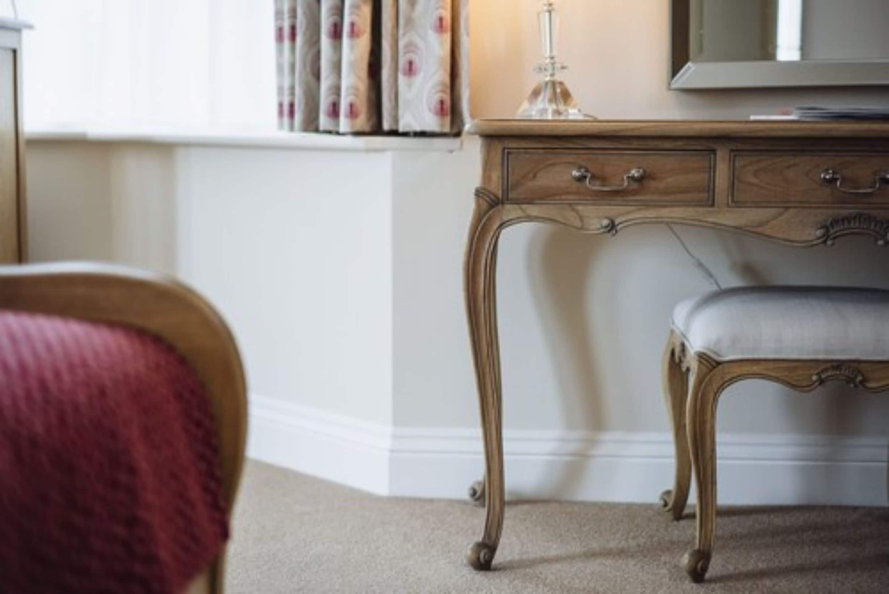 A wooden desk with drawers and a lamp, a chair with a cushion, and a window with curtains.