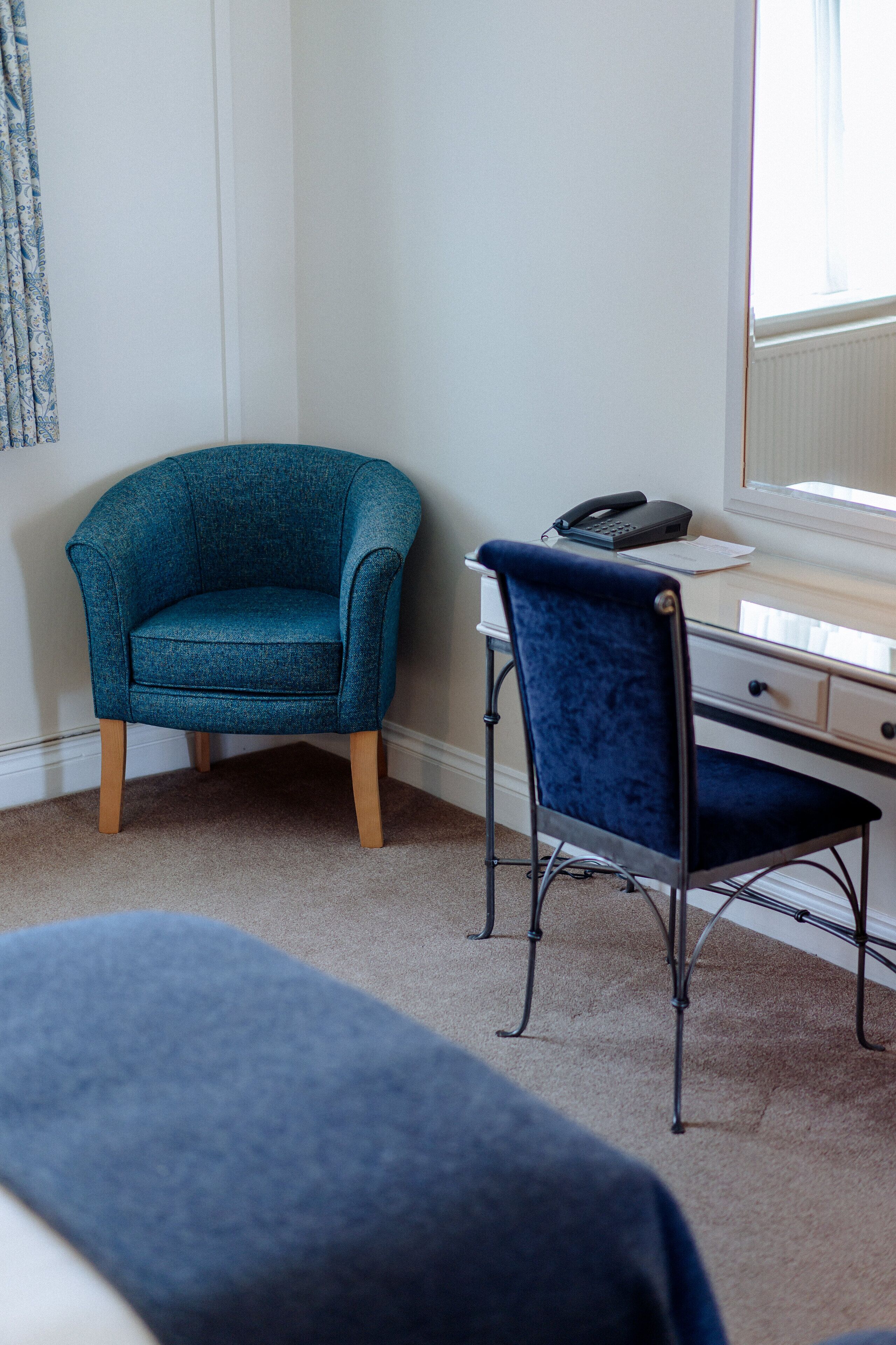 Een kamer met een smaragdgroene fauteuil, een bureau met stoel en een raam met zonwering.