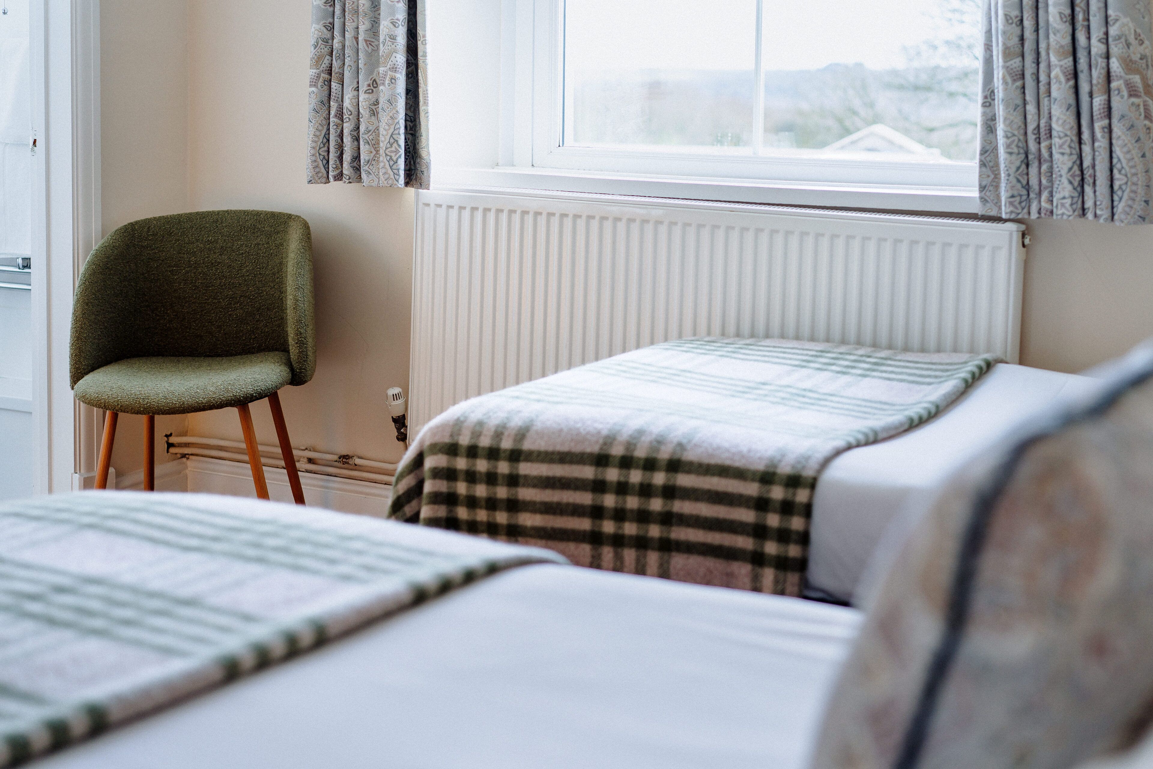 A bedroom with two beds, a chair, a radiator, and a window with curtains.