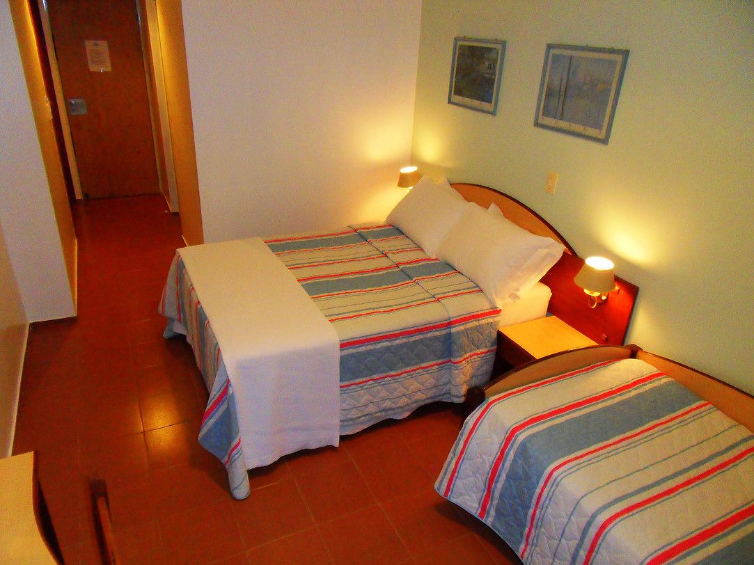 Una habitación de hotel con dos camas, cada una con ropa de cama a rayas y cabeceros blancos. Hay una mesita de noche con una lámpara entre las camas.