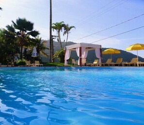 A swimming pool with lounge chairs and umbrellas, surrounded by palm trees and a mountain backdrop. - Hotel Torremolinos (Ubatuba)