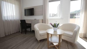 A modern hotel room with a wooden floor, a desk with a chair, a TV mounted on the wall, two white armchairs around a small round table with a vase of flowers, and large windows with curtains. - Hotel Europa (Marostica)