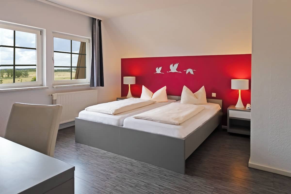 A hotel room with a large bed, two bedside tables, a chair, and a view of the outdoors through two windows.