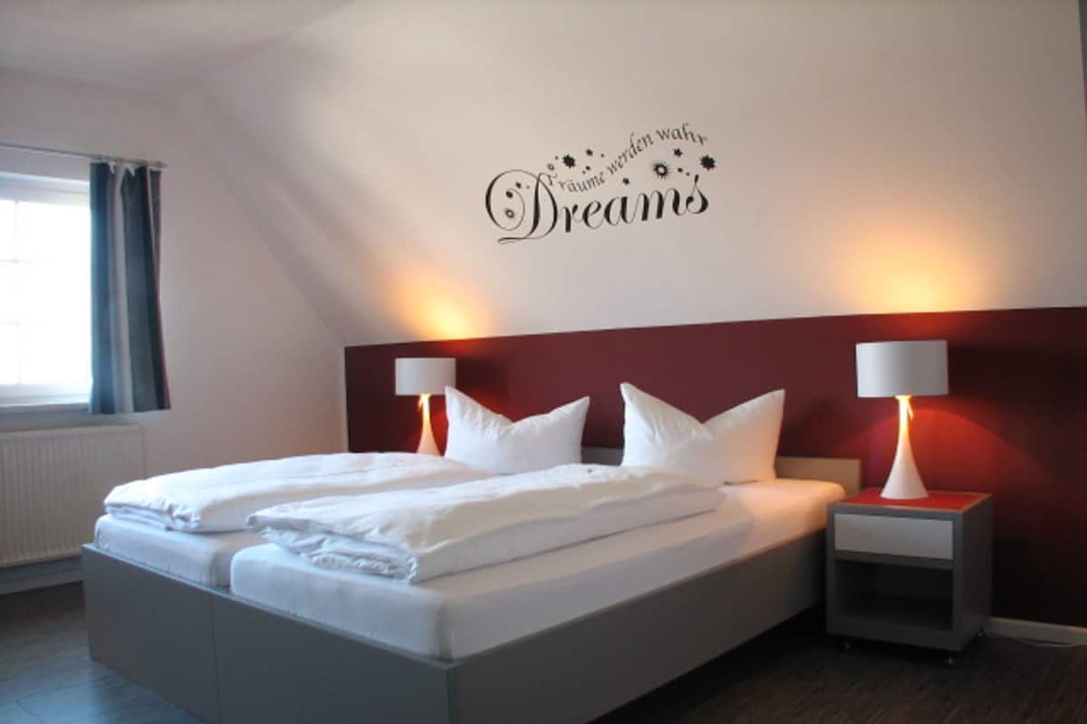 A modern hotel room with a large bed, two bedside tables with lamps, a red accent wall, and a decorative phrase on the wall.