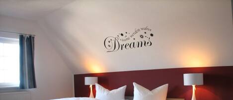 A modern hotel room with a large bed, two bedside tables with lamps, a red accent wall, and a decorative phrase on the wall.